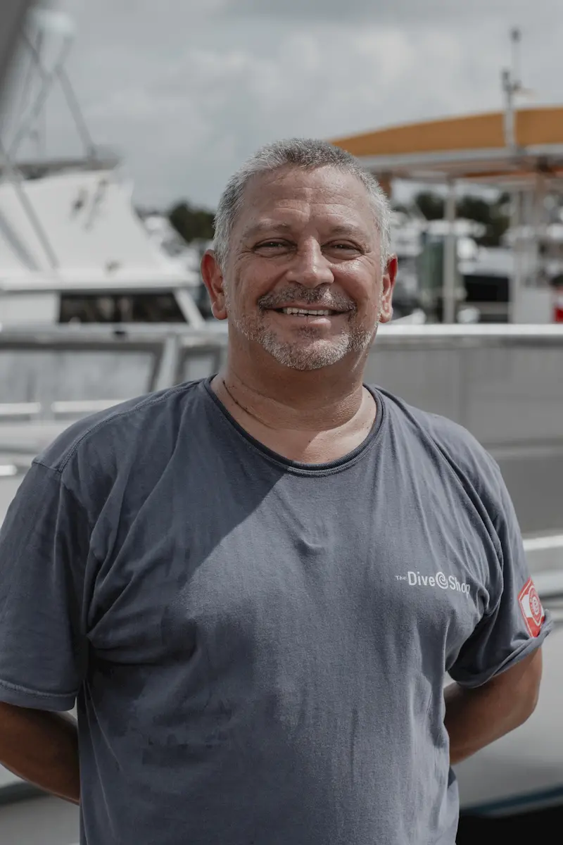 A smiling man with short gray hair and a beard stands outdoors in front of a docked boat, wearing a gray t-shirt with "Dive@Sqd" written on it, embracing the sea heritage. The background shows more boats and a cloudy sky.
