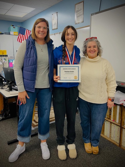 Three people stand smiling in a classroom. The person in the middle holds a certificate and wears a medal. The two adults on either side are casually dressed, and there’s an American flag in the background.
