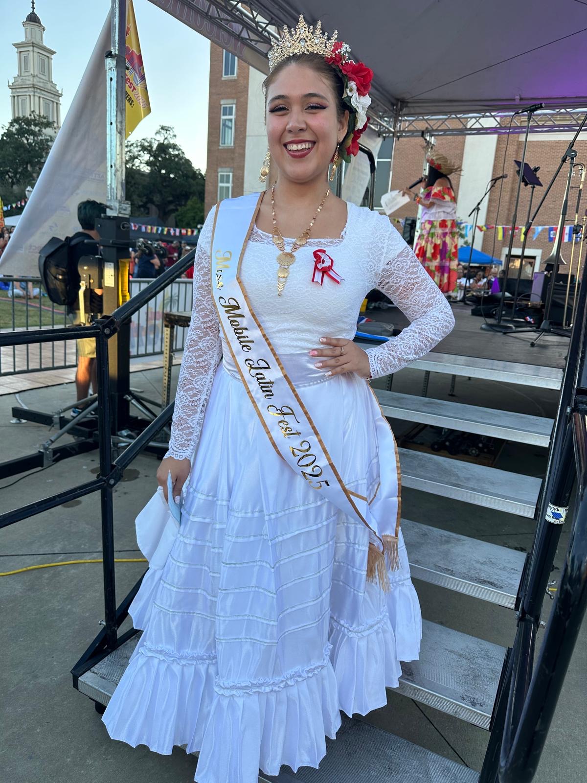 A smiling woman stands outdoors in a traditional white dress with lace sleeves, wearing a sash that reads "Srta. Idilio Tamu Fest 2023," a crown, and red and white ribbon, holding papers near a stage at a festival.