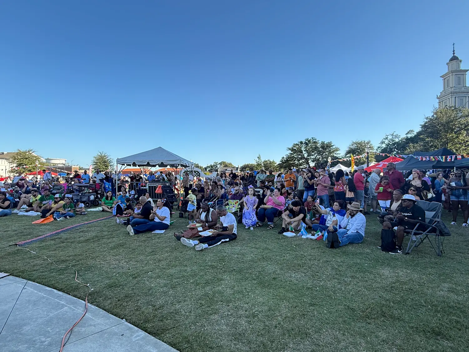 A large crowd of people sits and stands on a grassy lawn at an outdoor event, with tents and flags in the background under a clear blue sky. Some people are seated on blankets or chairs, enjoying the festivities.