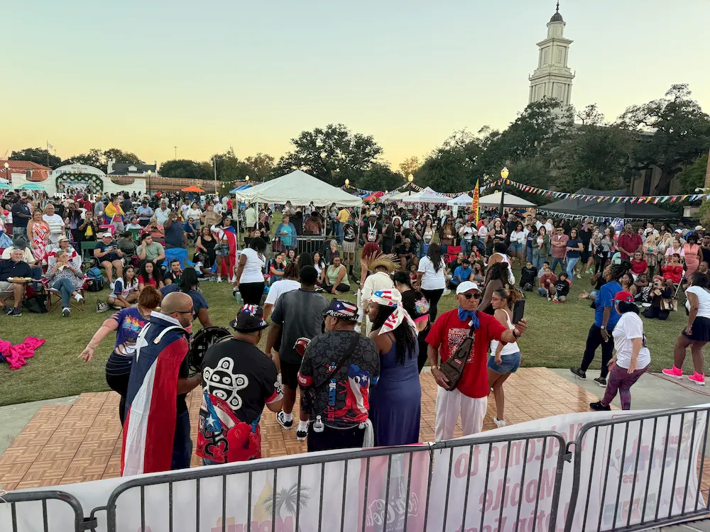 A large crowd gathers outdoors at a festival, with people dancing on a wooden floor in the foreground. Tents, decorations, and a tall white tower are visible in the background under a clear sky.