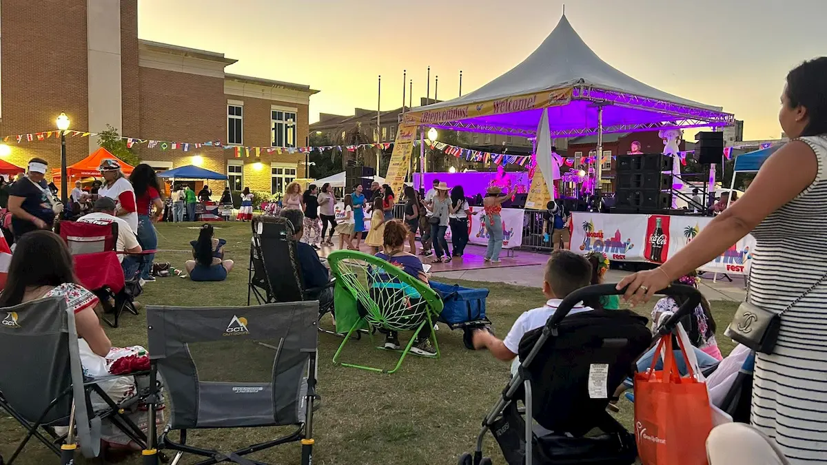 People attend an outdoor festival at sunset with tents, a lit stage, colorful flags, and live entertainment. Some sit on lawn chairs or grass, while others walk or push strollers, enjoying the festive atmosphere.