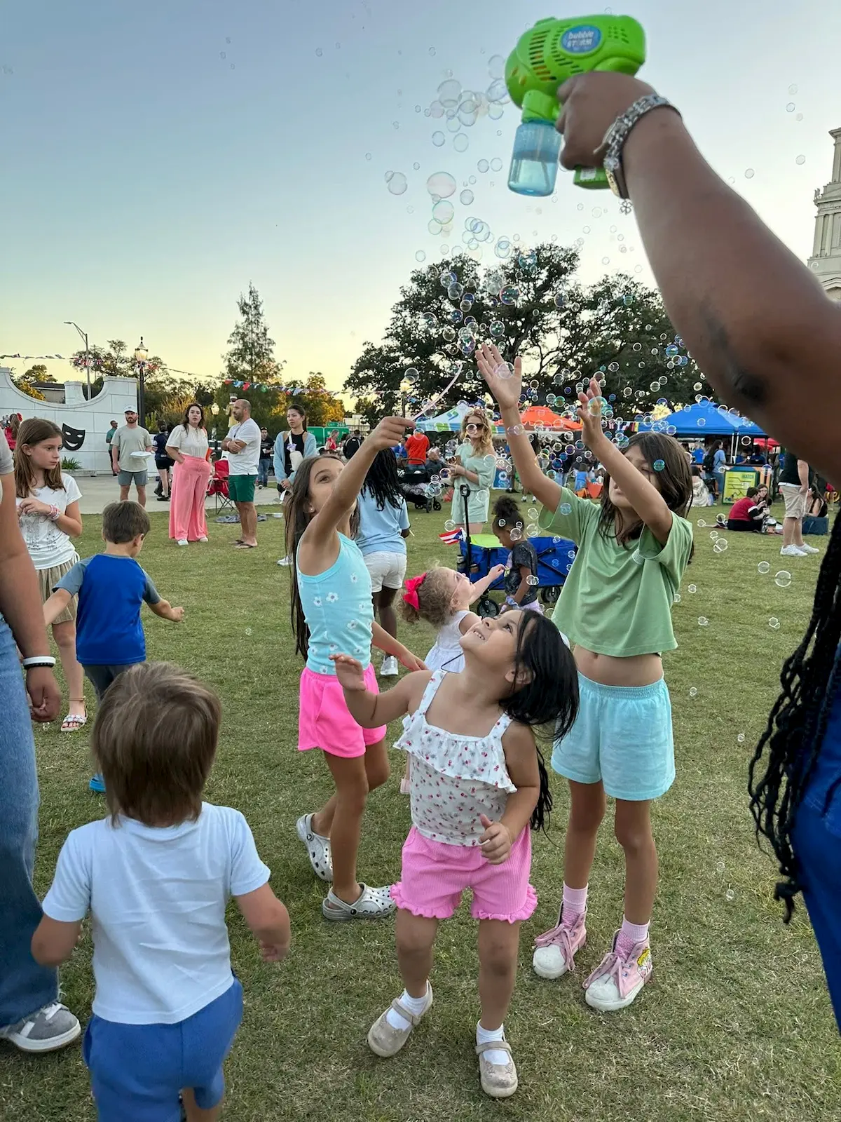 Children play on grass while someone blows bubbles with a bubble gun. Several kids reach up excitedly toward the bubbles. Adults and more children are visible in the background at an outdoor event or festival.