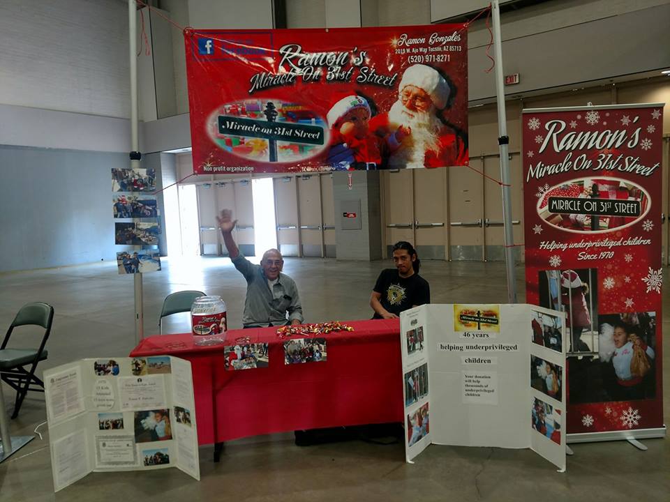 Two men sit at a decorated table under a banner for "Ramon’s Miracle On 3rd Street" at an indoor event. They are surrounded by informational boards and holiday decorations. One man is waving and smiling at the camera.
