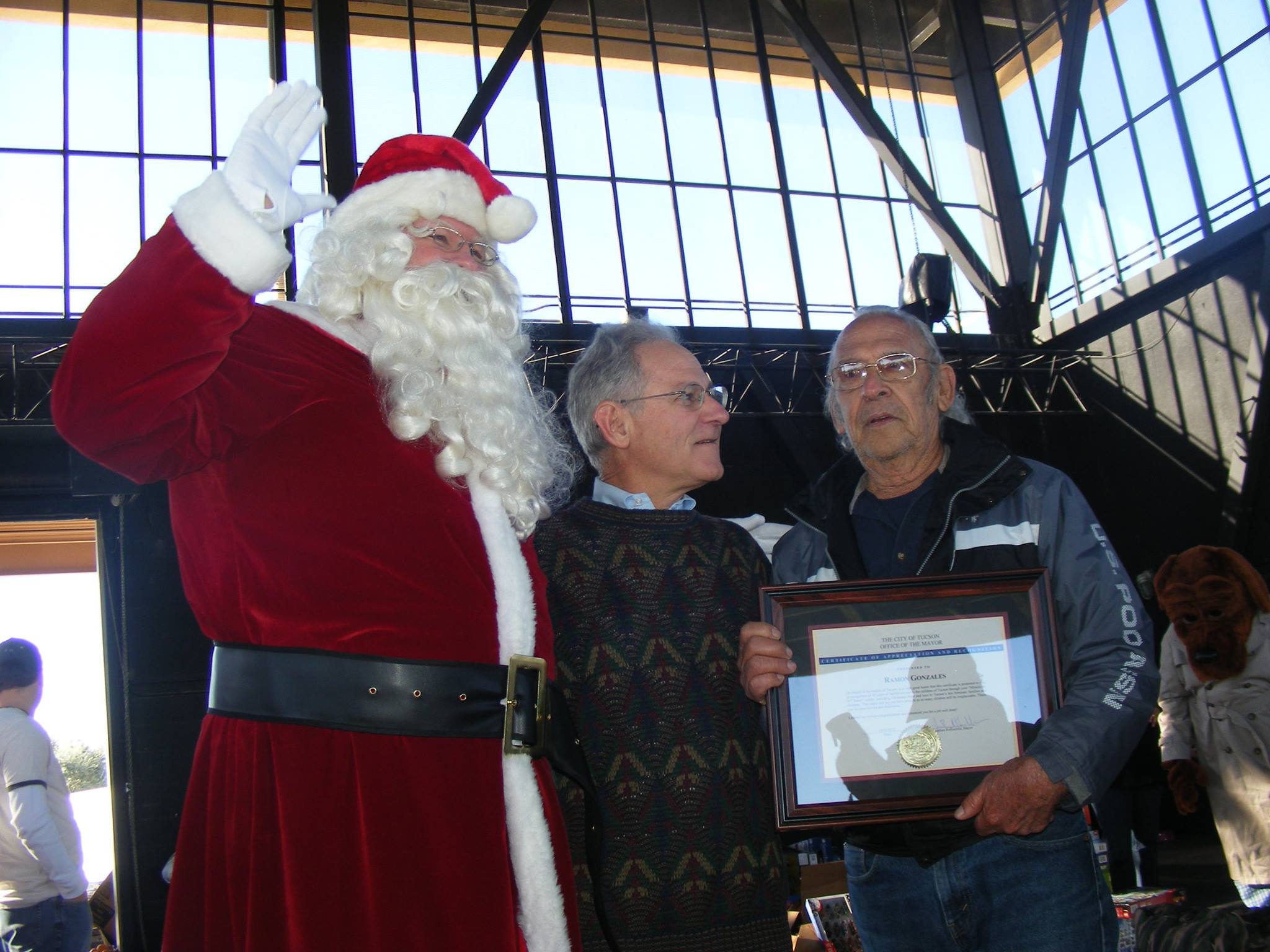 A person dressed as Santa Claus stands next to two men, one of whom is holding a framed certificate and a medal. They are indoors with sunlight streaming through large windows behind them.