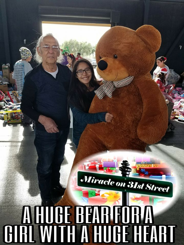 An elderly man and a smiling young woman stand indoors, hugging a giant teddy bear. Behind them are tables with colorful gifts. Text reads: “A HUGE BEAR FOR A GIRL WITH A HUGE HEART – Miracle on 31st Street.”.