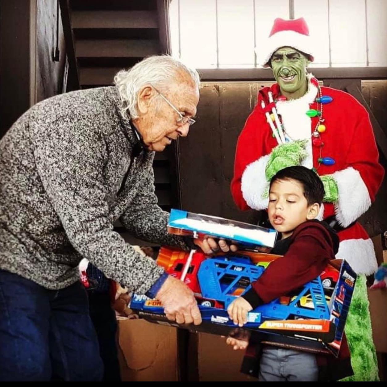 An elderly man helps a young boy hold several boxed toys while a person dressed as the Grinch in a Santa outfit stands smiling behind them.