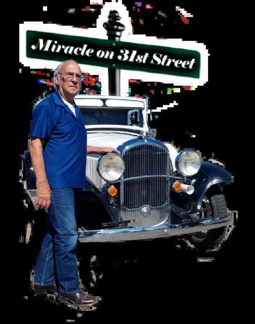 An older man in a blue shirt and jeans stands beside a vintage navy and white car, with a “Miracle on 31st Street” street sign visible in the background.