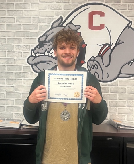 A smiling person stands indoors in front of a bulldog mural and holds a Sunshine State Scholar certificate. They are wearing a medal and a green jacket over a t-shirt.