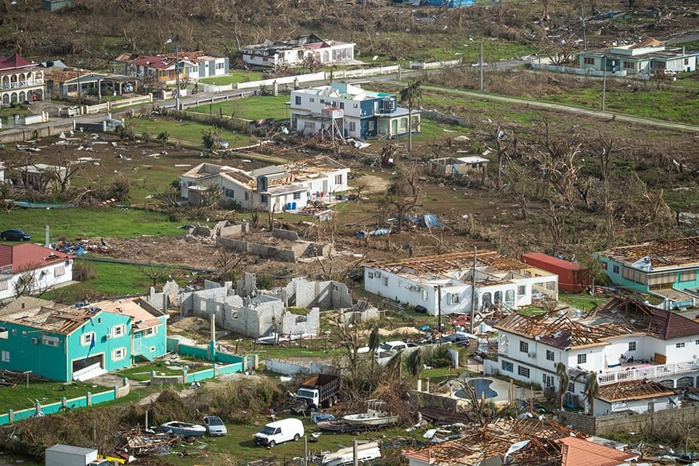 Aerial view of a residential area with many houses damaged or destroyed, roofs missing, and debris scattered after a natural disaster, likely a hurricane. The landscape appears devastated and barren.
