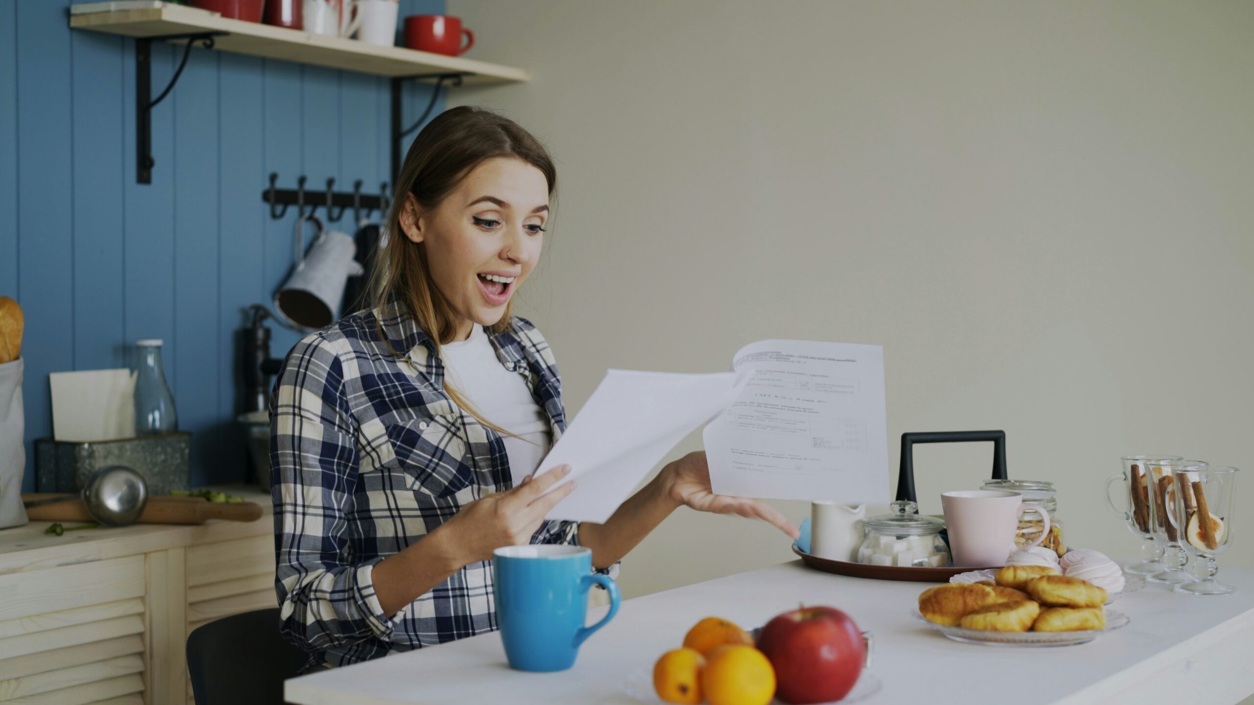 A young woman sits at a kitchen table, smiling excitedly while holding and looking at paperwork. Surrounded by a blue mug, fruits, a croissant, and a pink bowl, she embraces positive habits that support her emotional health. Kitchen shelves are visible behind her.