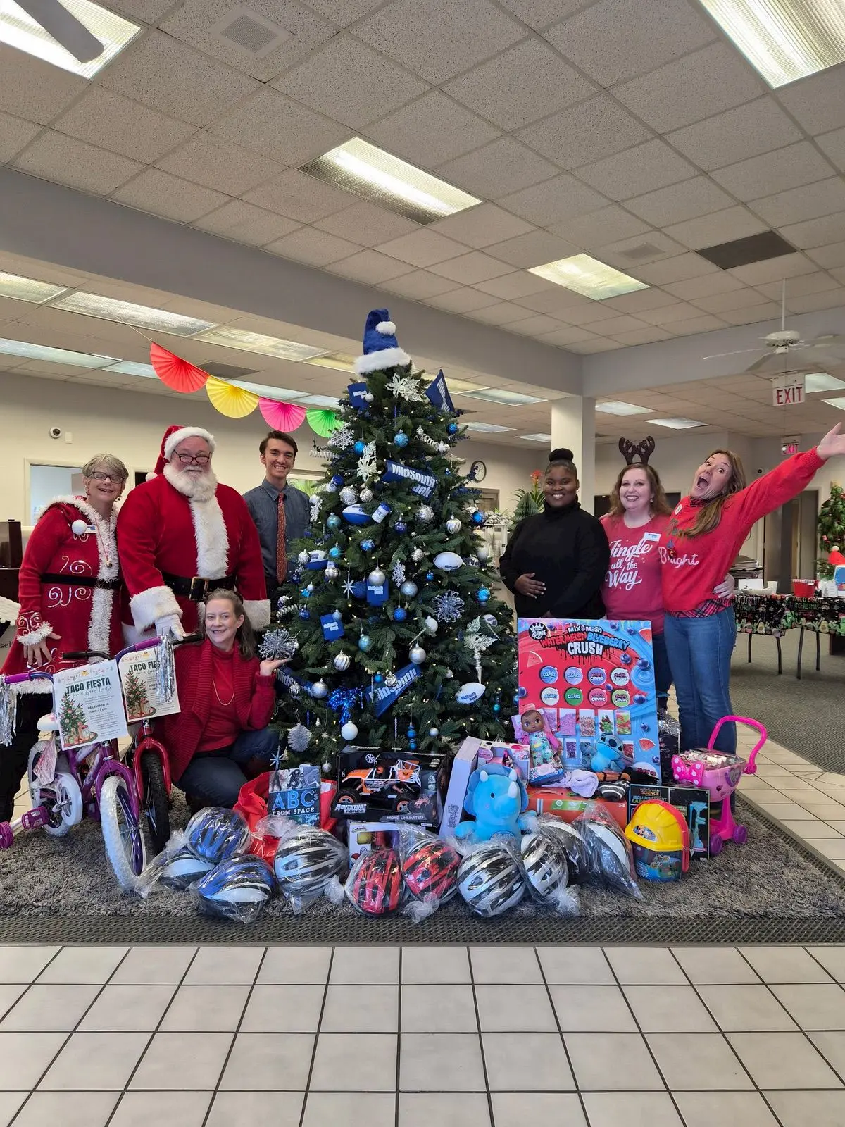 A group of people, including someone dressed as Santa, stand and kneel around a decorated Christmas tree with gifts and toys underneath. They are smiling and posing cheerfully in a festive indoor setting.