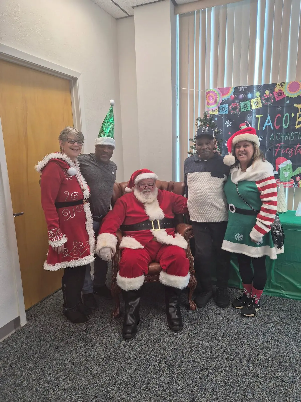 Five people pose indoors; one dressed as Santa sits in a chair, while the others—wearing festive holiday outfits and hats—stand around him, smiling. A green holiday-themed backdrop is behind them.