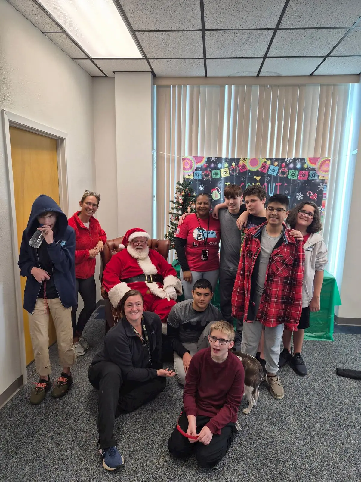 A group of students and adults pose indoors with Santa Claus in front of a decorated Christmas tree and festive backdrop. Most people are smiling and wearing casual or holiday-themed clothes.