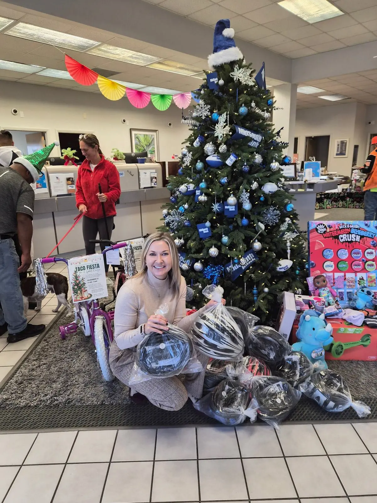 A smiling woman kneels beside a Christmas tree decorated with blue ornaments, holding a black bicycle helmet. Gifts, including helmets, toys, and a bicycle, are arranged around the tree. Other people stand in the background.