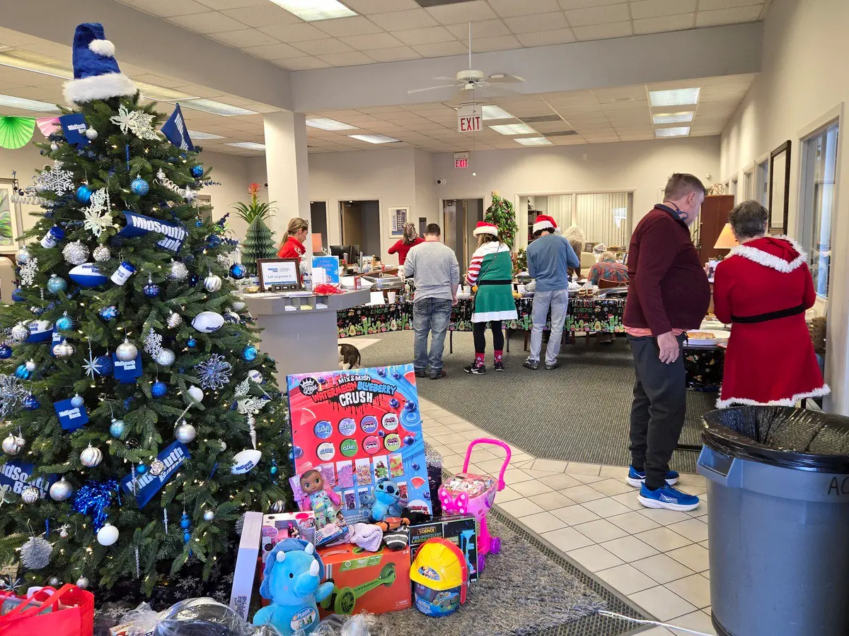 A decorated Christmas tree stands near tables filled with toys and gifts. People, some dressed in festive holiday outfits, are shopping or organizing items in a brightly lit indoor space.