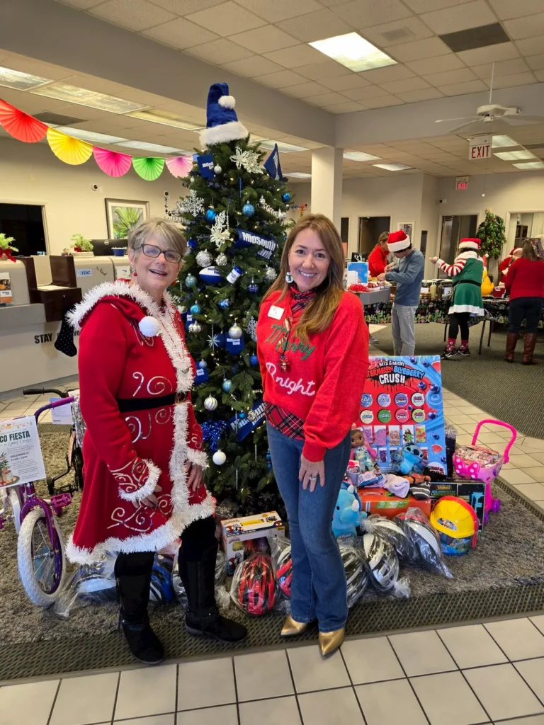Two women in festive holiday sweaters stand in front of a decorated Christmas tree and a collection of toys and gifts, inside a brightly lit room with more people and decorations in the background.