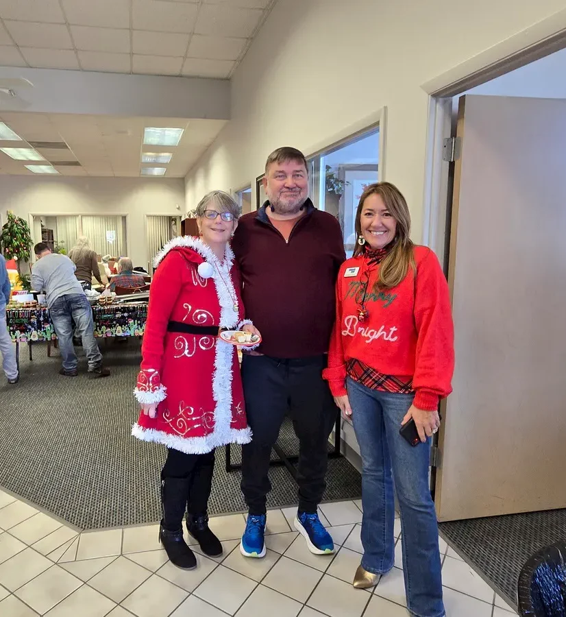 Three adults pose and smile at a holiday gathering; one woman wears a festive red Santa coat, the man wears a maroon sweater, and the other woman wears a red “Bright” sweater. People and holiday decorations are visible in the background.