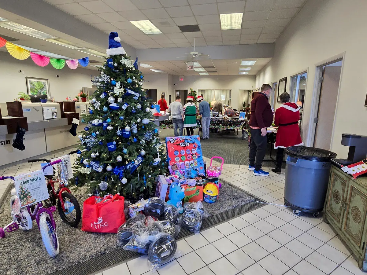 A decorated Christmas tree stands indoors with gifts, toys, and bikes at its base. People, some dressed in festive costumes, gather around tables in the background, celebrating a holiday event.