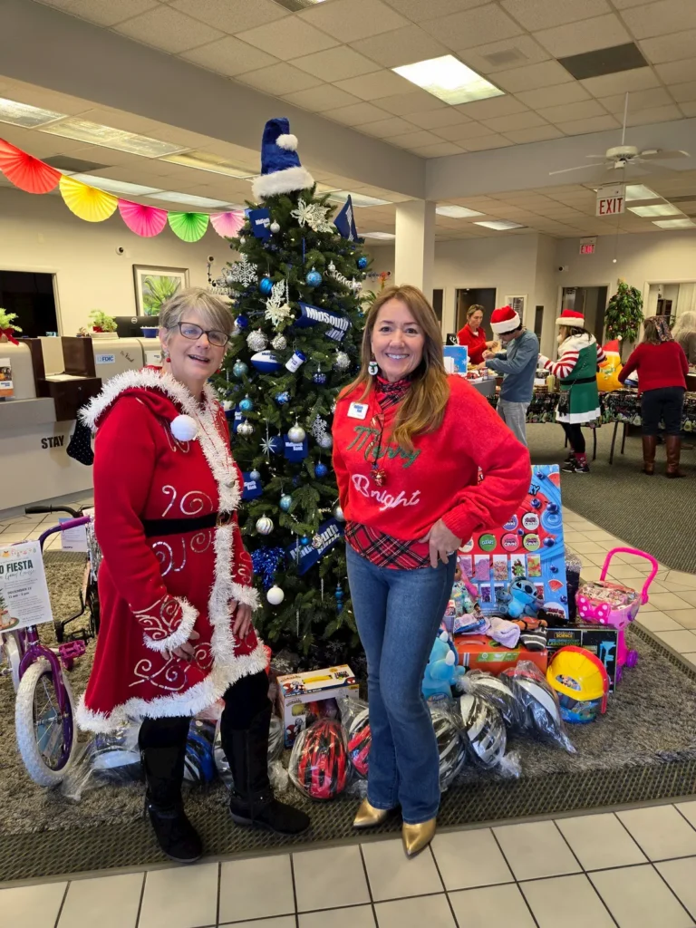Two women in festive holiday outfits pose in front of a decorated Christmas tree with gifts and toys beneath it. Other people, holiday decorations, and an office setting are visible in the background.