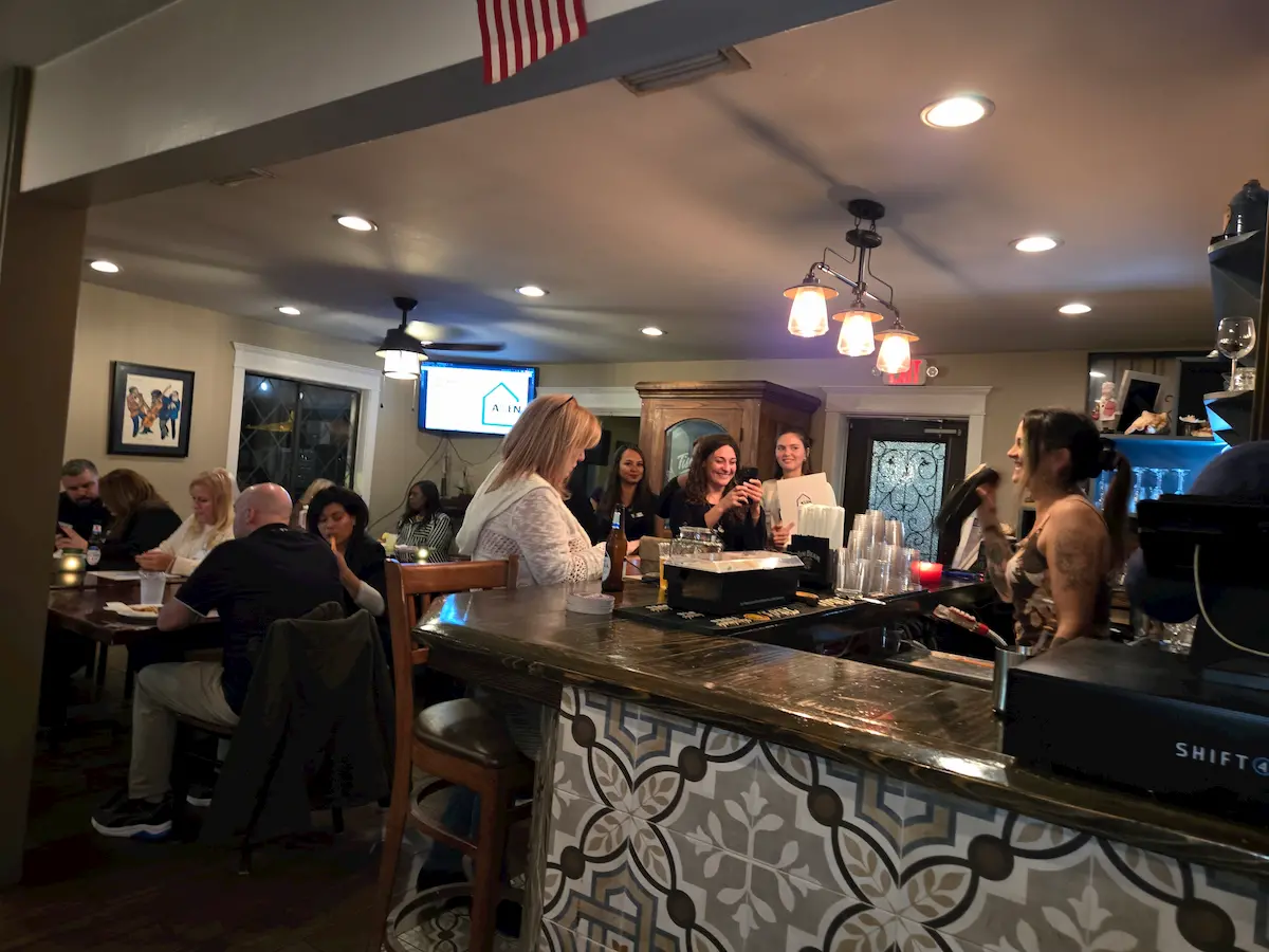 People sit and socialize at a bar and restaurant. Some are seated at the counter talking to the bartender, while others dine at tables. The atmosphere is lively and warm, with soft lighting and patterned tile decor.