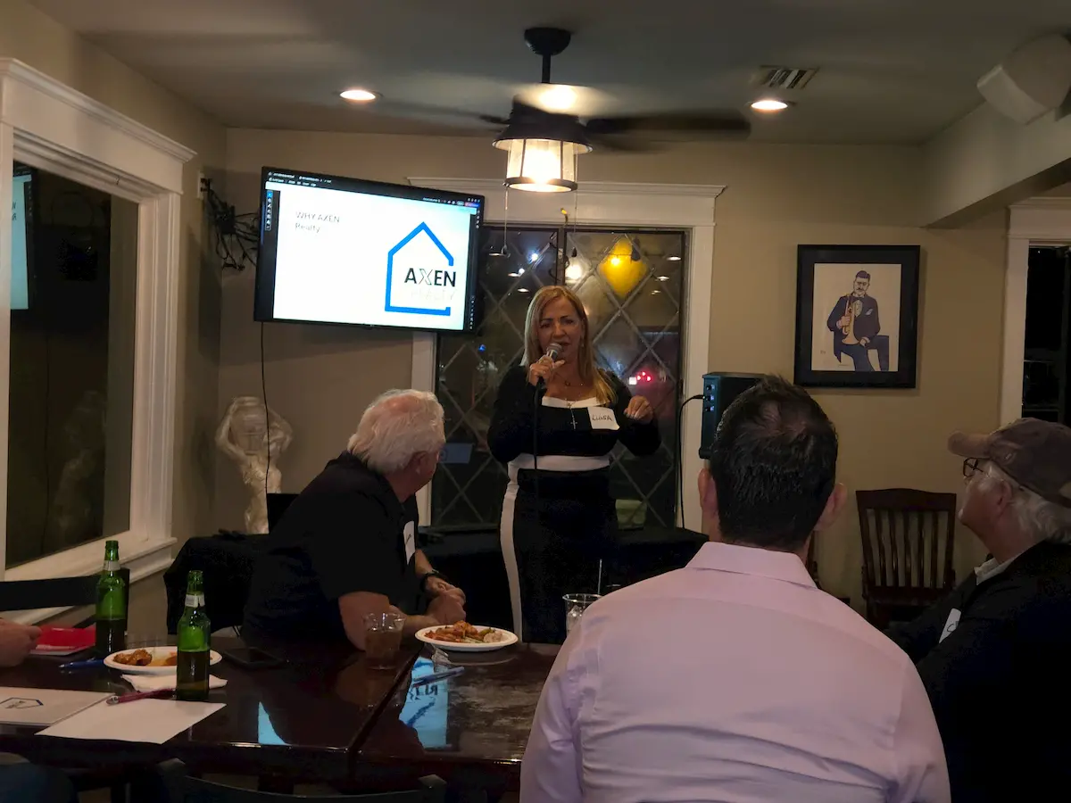 A woman speaks into a microphone in a small room, addressing four seated people. A presentation screen behind her displays the Axen Realty logo. There are drinks and plates of food on the table.