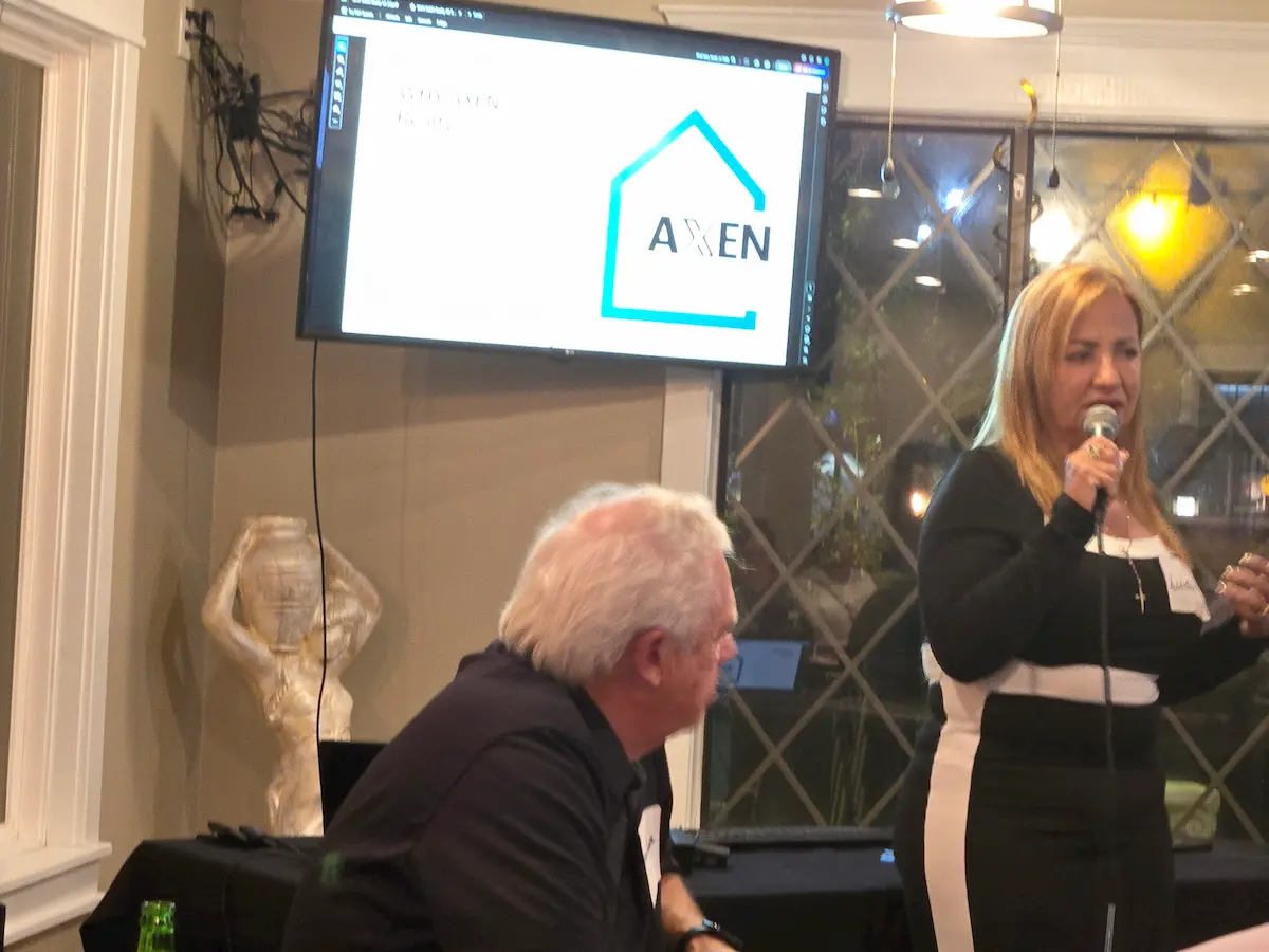 A woman speaks into a microphone while standing next to a seated man at an indoor event. A screen behind them displays a presentation with the Axen logo and some text. A sculptural figure and window are visible in the background.