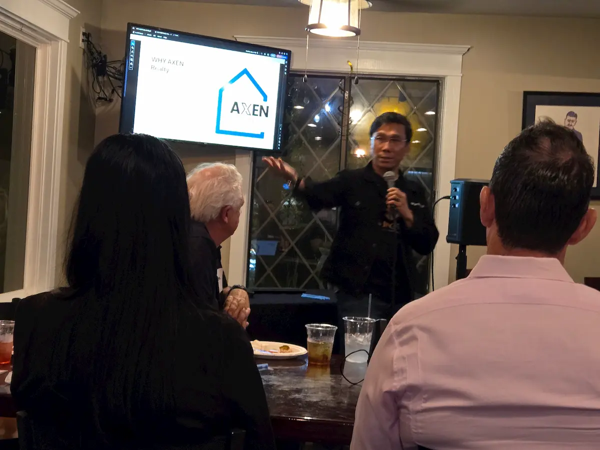 A person gives a presentation using a microphone in a restaurant. Three people sit at a table watching, with drinks and plates in front of them. A screen displays a slide with the word "AXEN" and a house logo.