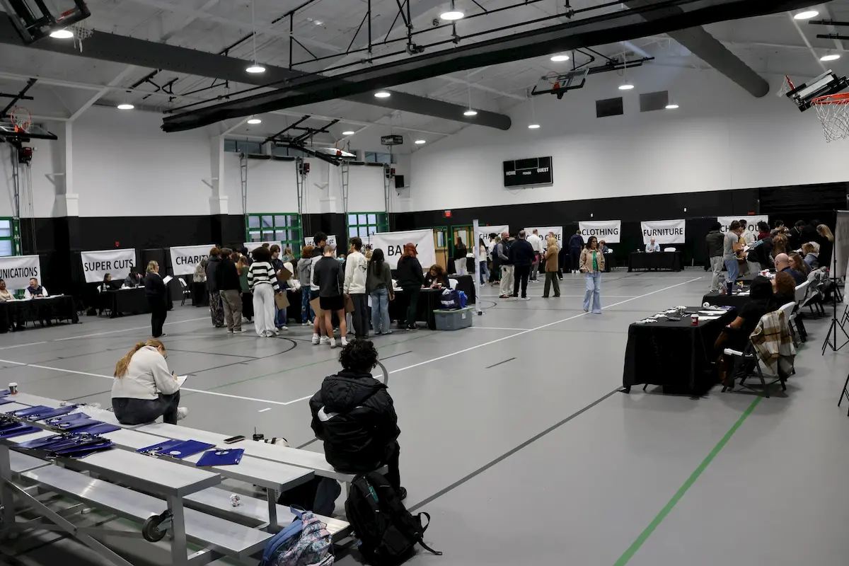 People gather in a gymnasium at multiple booths for a job fair or event. Signs on booths include "COMMUNICATIONS," "SUPER CUTS," "CHARTER," and "CLOTHING." Some individuals are seated while others interact at booths.