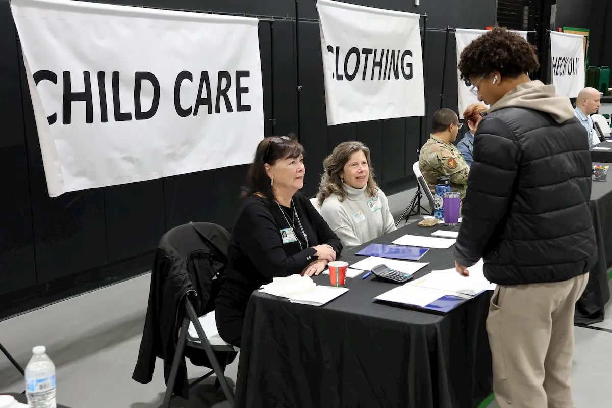 Two women sit at a table labeled "CHILD CARE," speaking with a person standing in front of them. Behind them are large banners that read "CHILD CARE" and "CLOTHING." Other tables and people are visible in the background.
