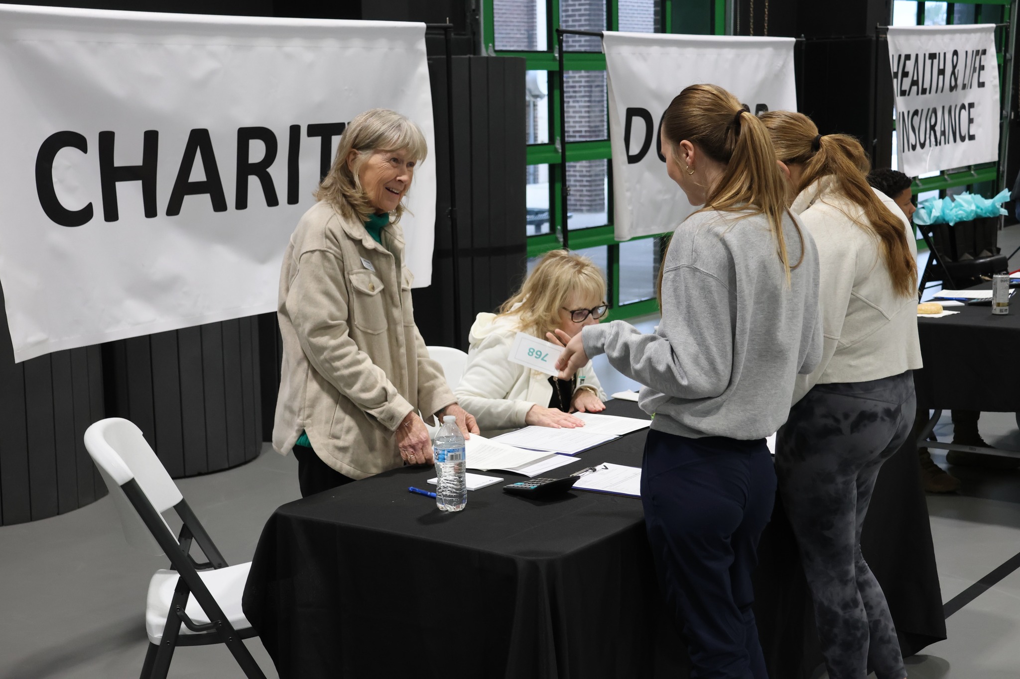 Two women sit behind a table labeled “CHARITY,” talking with three standing women who hold papers. Other booths with signs like “HEALTH & LIFE INSURANCE” are visible in the background.