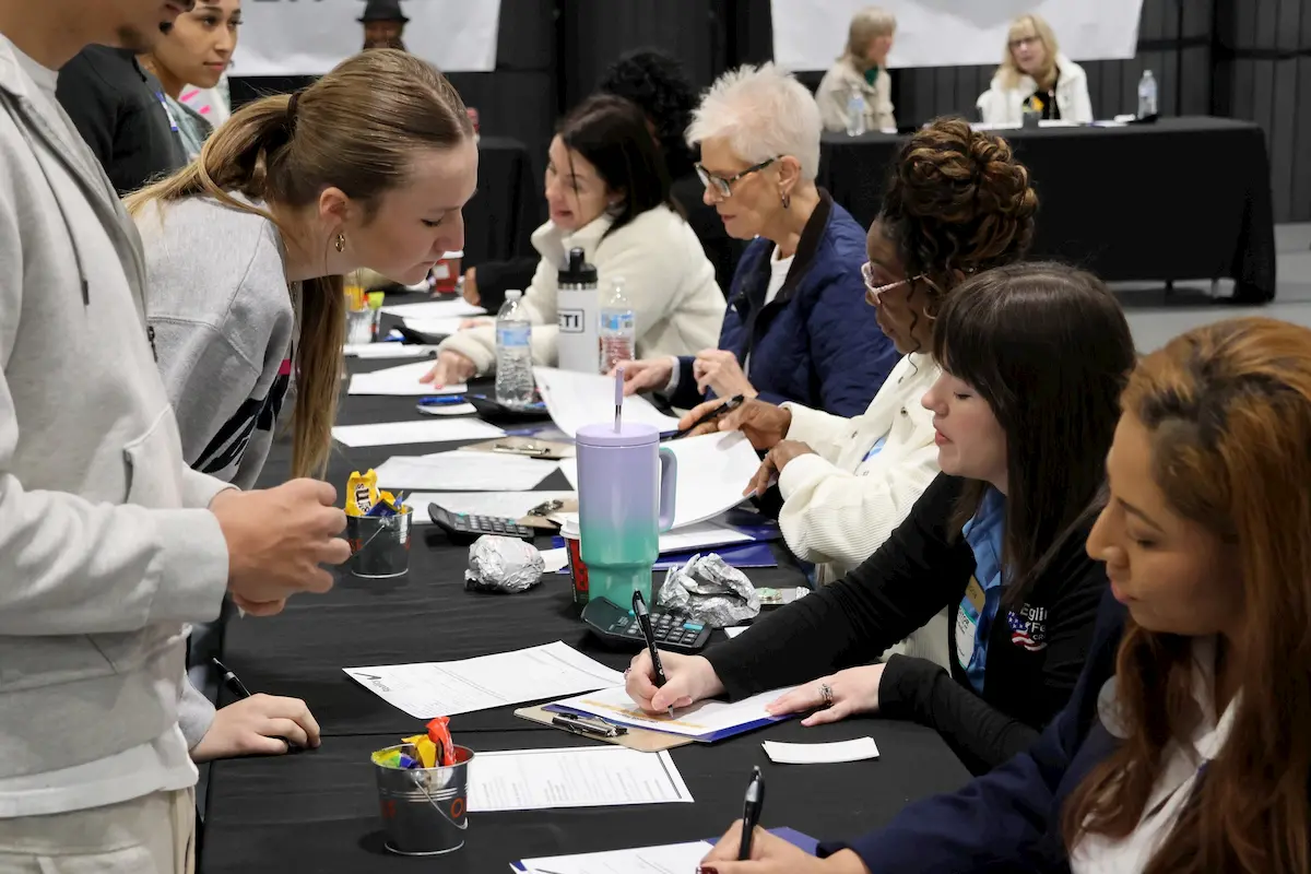 Several people sit at a long table covered with papers and clipboards, assisting others who are standing and filling out forms, in a busy indoor setting.