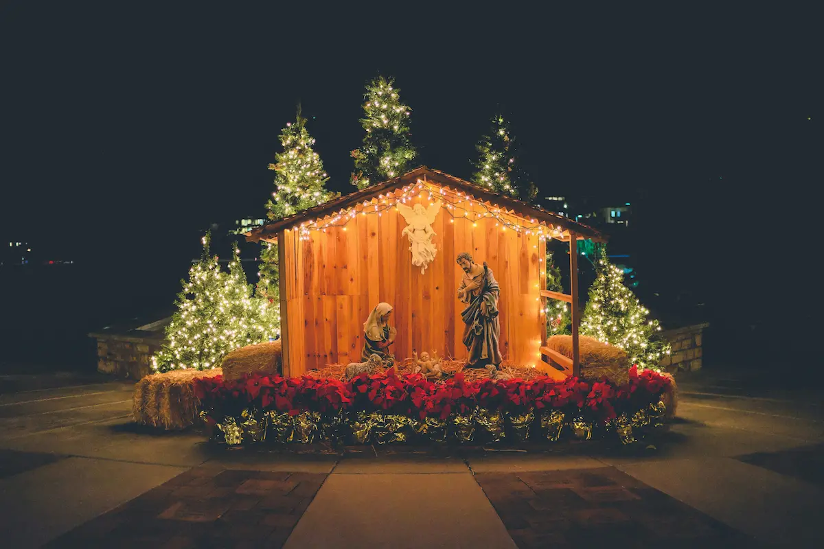 Outdoor nativity scene with Mary, Joseph, and baby Jesus under a lit wooden shelter, surrounded by glowing Christmas trees and red poinsettias, inspiring all to fall on our knees in awe this Christmas night.