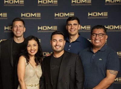 Five people, three men standing in the back and a man and woman in front, smile at the camera in front of a black backdrop with gold "HOME" logos.