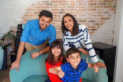 A smiling family of four poses together indoors. Two adults stand behind a green sofa, while two children sit on it. The background features a white brick wall and a leafy plant.