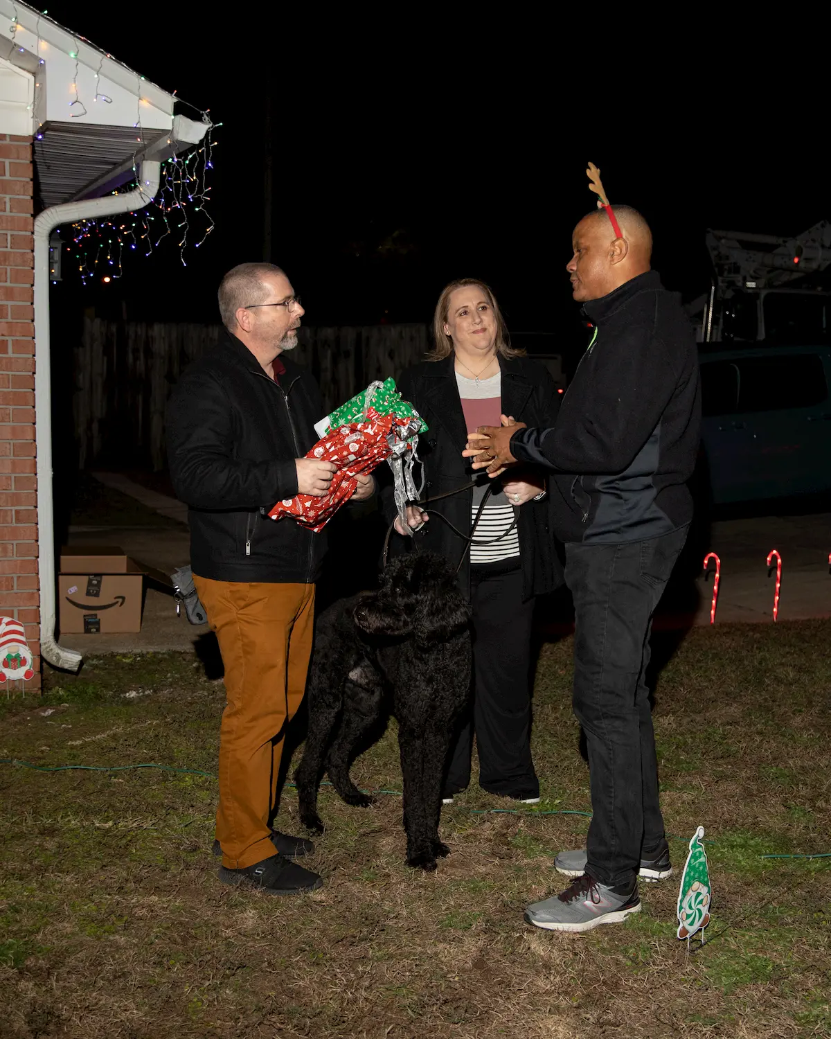 Three adults stand outside at night, talking and holding gifts and drinks. One wears reindeer antlers, a black dog stands between them, and FPL holiday decorations and lights twinkle in the background.