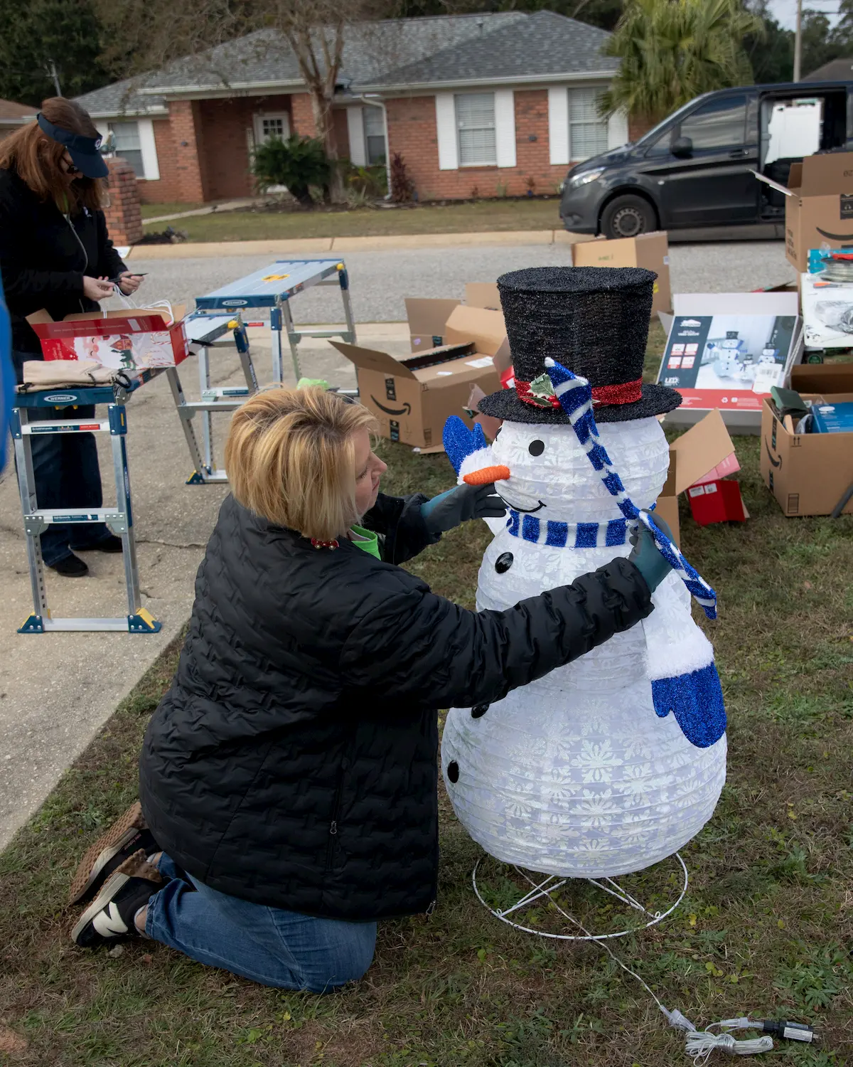 A person kneels on grass, assembling a light-up snowman decoration near open boxes and tools, as another stands nearby. The festive scene unfolds on a residential street with brick houses, parked cars, and support from FPL in the background.