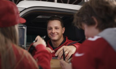 A man sitting in a car smiles as he signs autographs for fans wearing red sports jerseys, reaching out through the window.