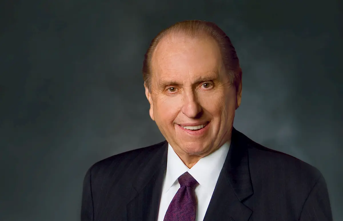 An older man with short, slicked-back hair smiles while wearing a dark suit, white shirt, and purple tie against a dark, neutral background.
