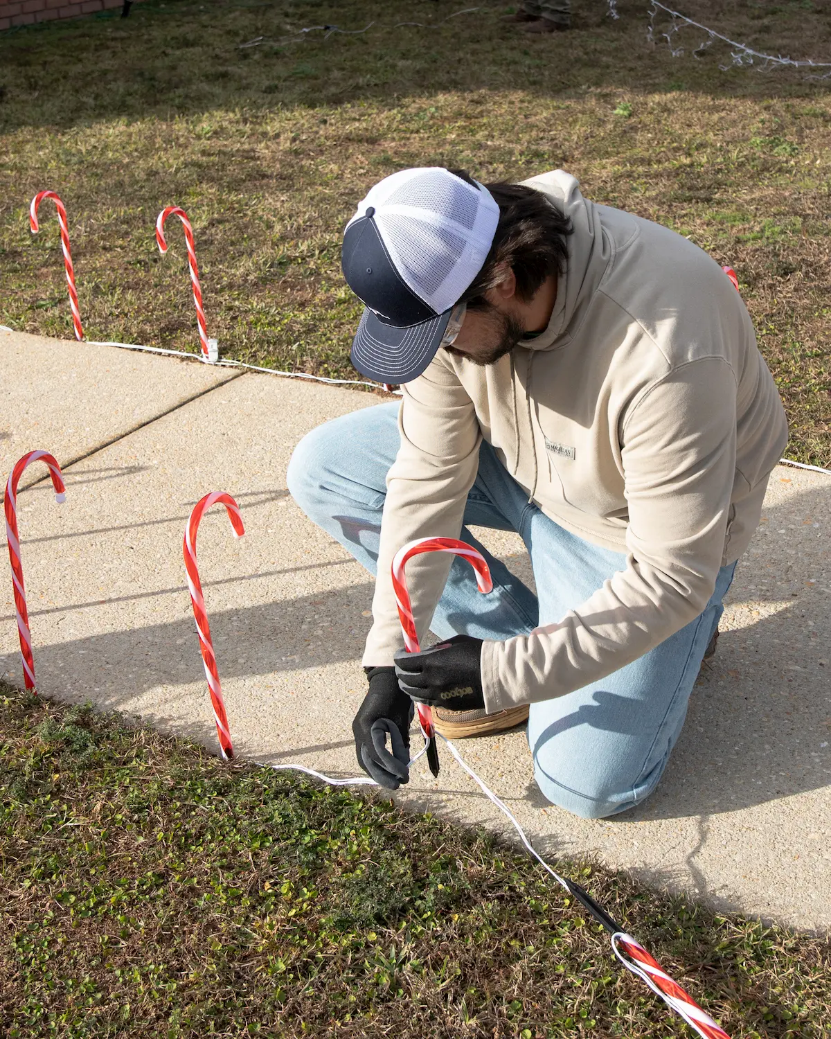 A person kneels on the ground to decorate the edge of a sidewalk with large candy cane decorations outside the Pensacola Veteran's Home. The person, dressed in a hoodie, jeans, gloves, and a cap, is surrounded by grass and more candy canes.