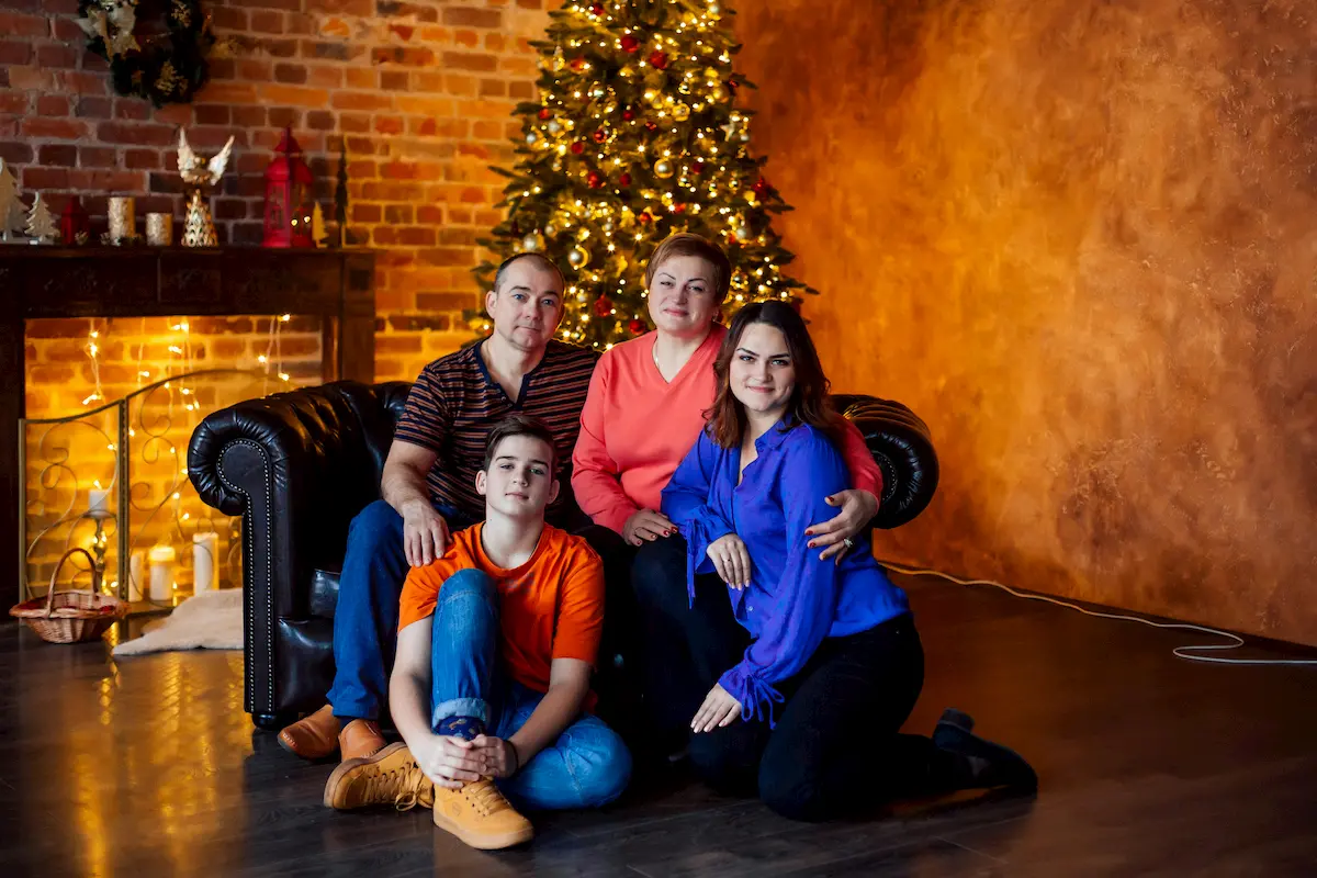 A united family of four poses together indoors in front of a decorated Christmas tree. Two adults sit on a couch while two young adults sit and kneel on the floor. The room is warmly lit with festive decorations and lights.