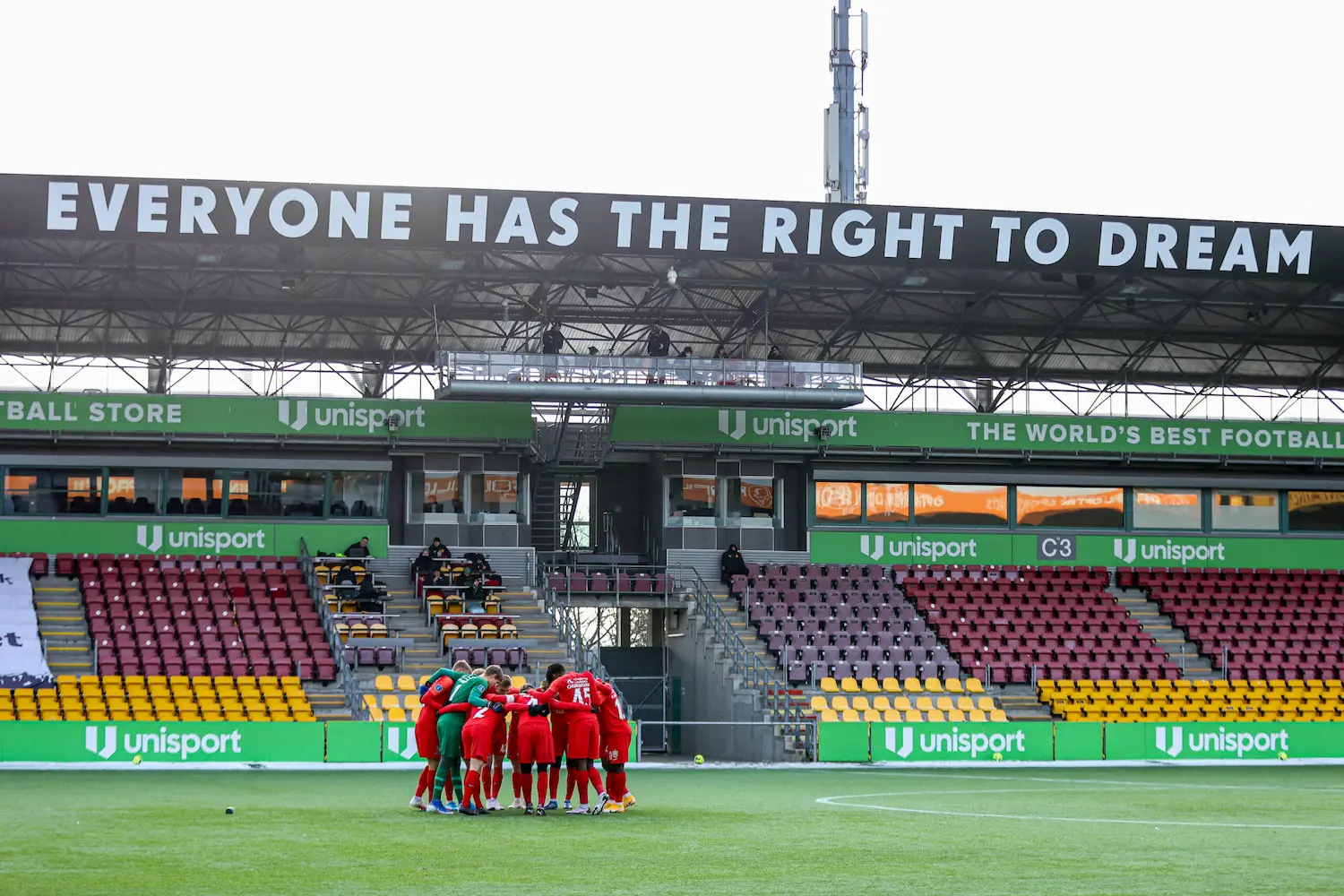 A soccer team in red uniforms huddles on the field in a mostly empty stadium. Above them, a large sign reads "EVERYONE HAS THE RIGHT TO DREAM." Green Unisport ads are visible in the stands.