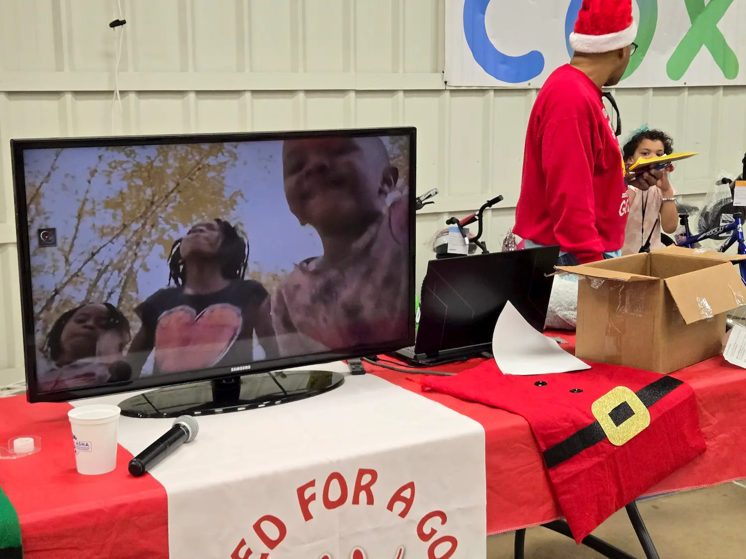 A television on a red table displays a video of smiling children. Next to it, a person in a Santa hat stands by an open box, and a child holds a toy. Holiday decorations and a microphone are visible on the table.