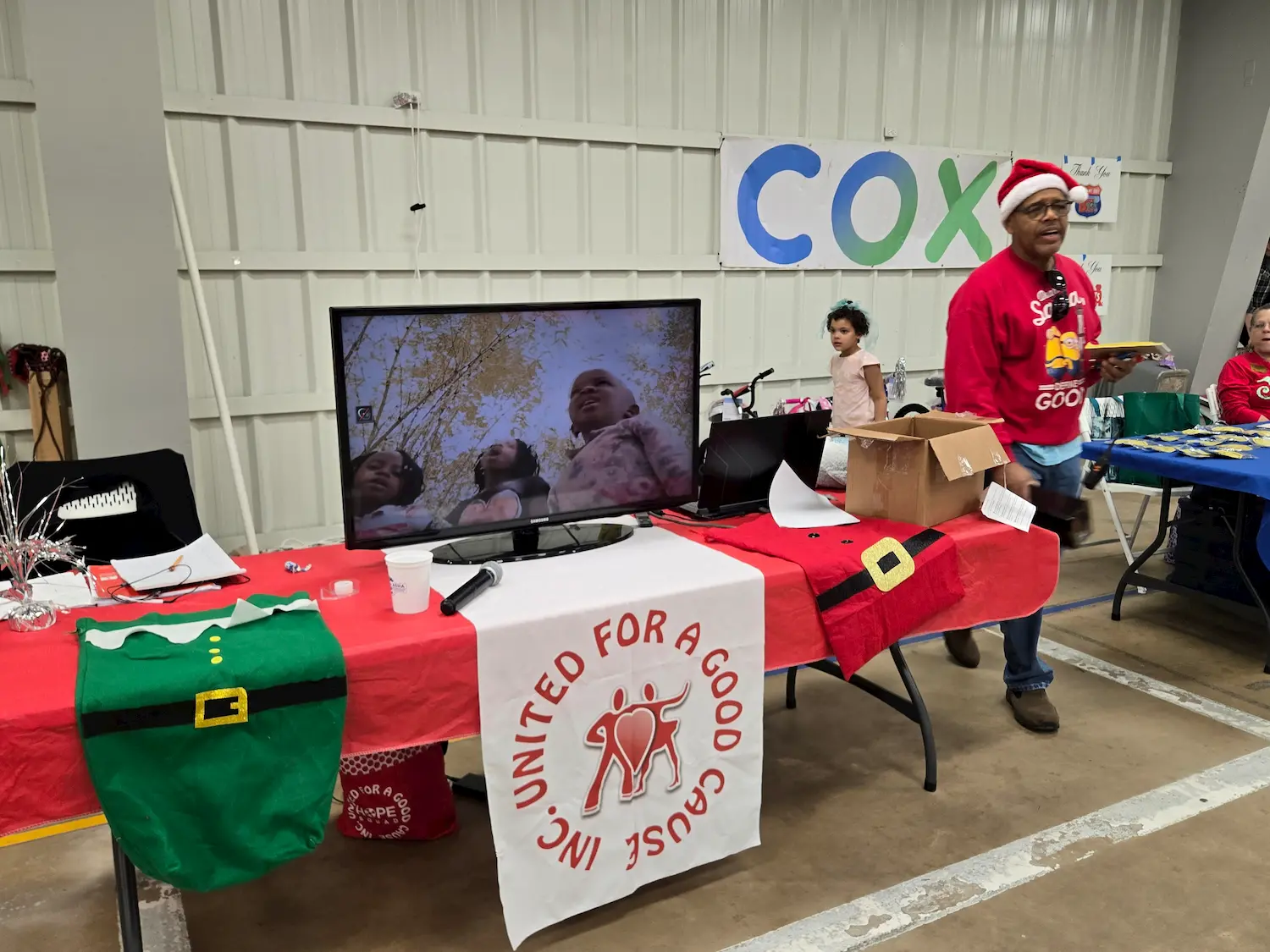 A volunteer in a Santa hat stands at a holiday-themed booth with a red tablecloth, TV, and “United For A Good Cause” banner. A child and another vendor are visible in the background at an indoor event.