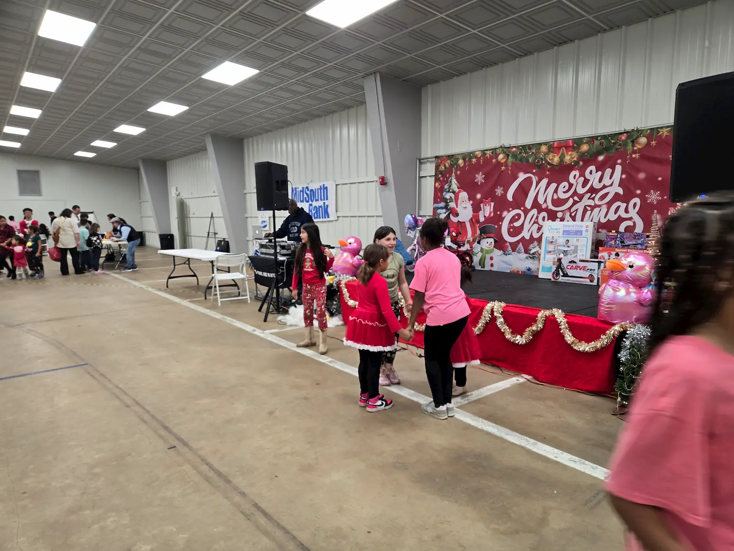 Children stand in line near a decorated stage with a "Merry Christmas" banner and holiday decorations, while others gather at tables in a large indoor event space.