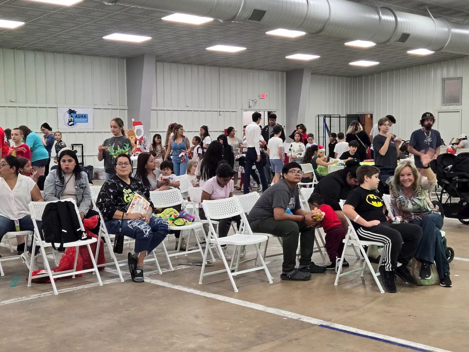 A diverse group of people, including children and adults, sit and stand in a large indoor gymnasium with white folding chairs. Some people are chatting, while others look toward the camera. The atmosphere appears lively and social.
