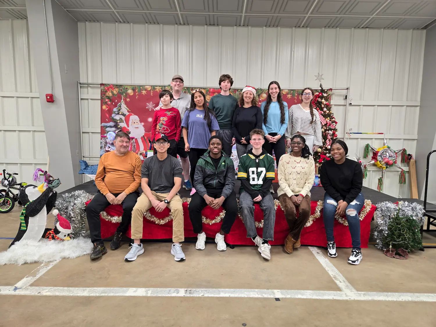 A group of thirteen people, some wearing festive clothing, pose for a photo on a stage decorated with holiday ornaments, a Christmas tree, and a Santa backdrop. Bicycles are visible to the left.