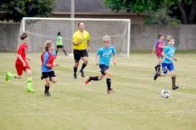 Children play a soccer game on a grassy field, with a referee and a goalpost in the background. The players, wearing different colored uniforms, are running toward the ball during the Youth Spring Soccer League.