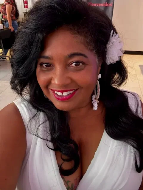 A woman with long black hair, red lipstick, white feather hair accessory, and pearl earrings smiles at the camera while wearing a white top.