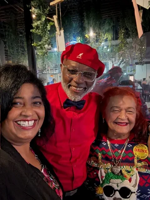 Three people smiling for a selfie indoors. The man in the center wears a red hat, glasses, and shirt with a bow tie. The two women beside him wear colorful clothing, and the background features plants and lights.